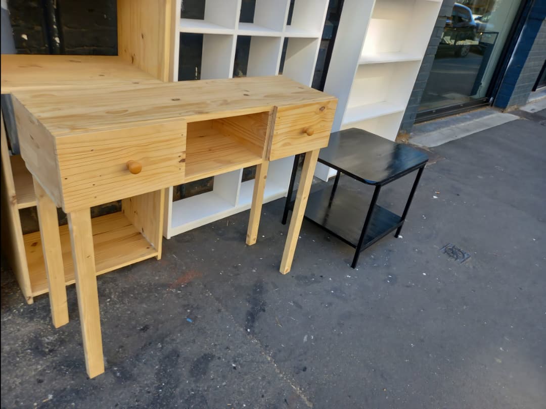 Un bureau en bois (ou une table de travail) avec des tiroirs, adossé à une grande étagère/bibliothèque blanche en arrière-plan.