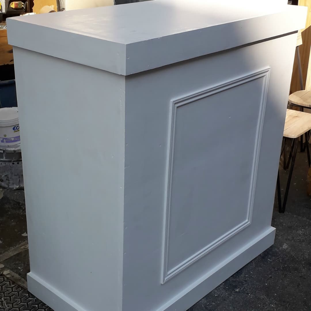 Un comptoir d'accueil (ou meuble de caisse / pupitre de réception) blanc ou gris clair avec des moulures rectangulaires sur la face avant.